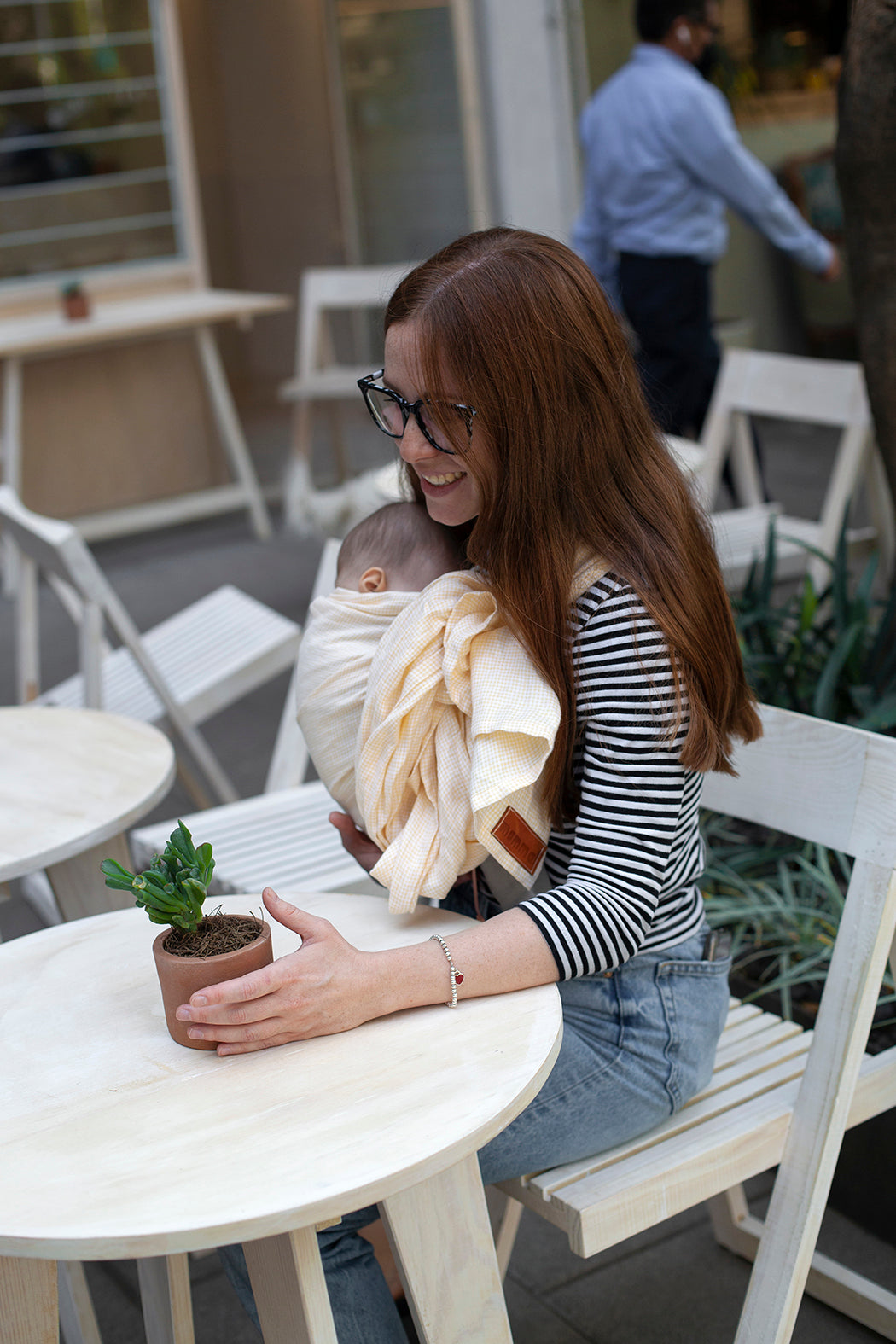 Mujer sentada en un café, cargando a su bebé en rebozo o bandolera de lino de anillos