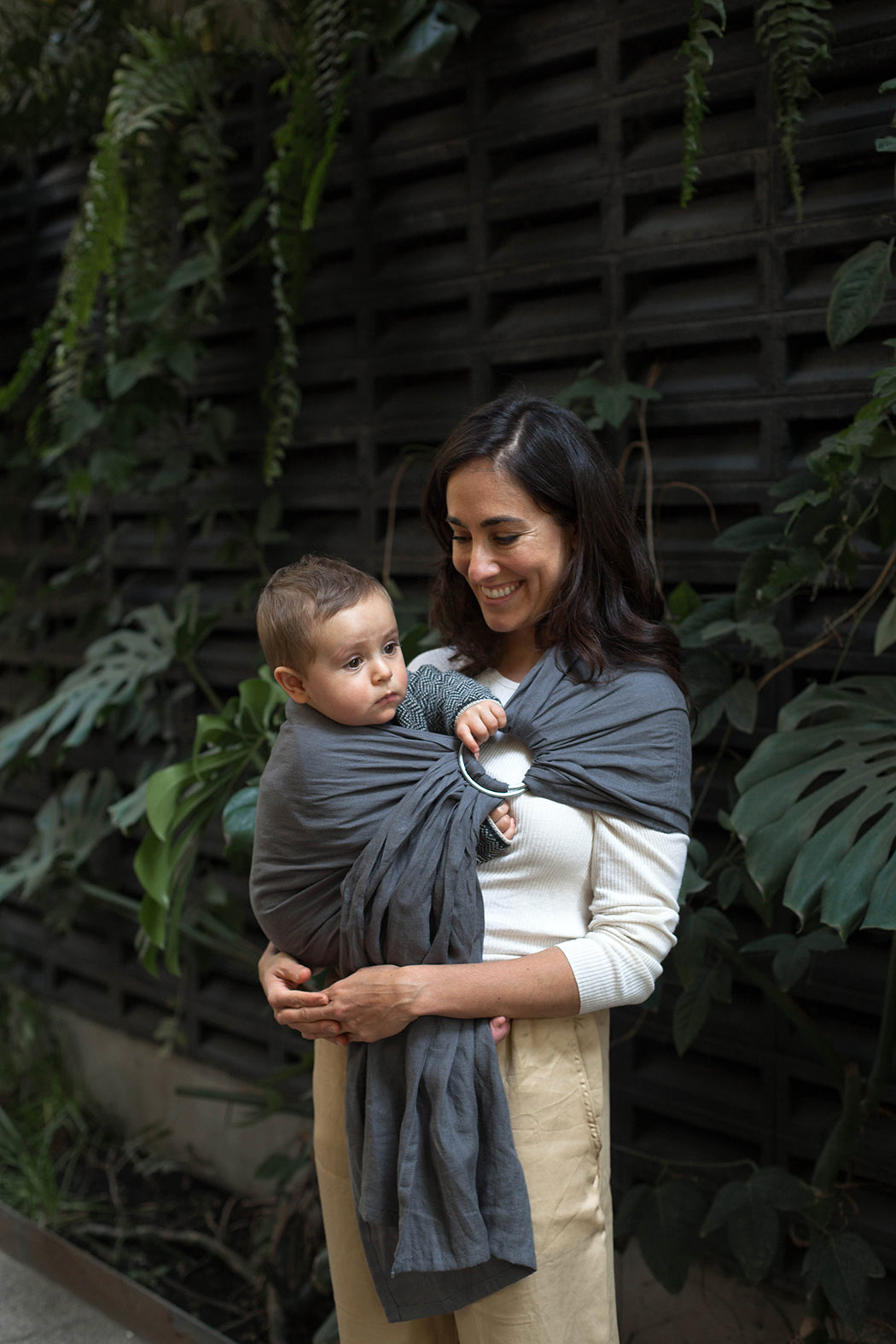 Mujer cargando a niño en rebozo o bandolera de anillas de lino con plantas al fondo